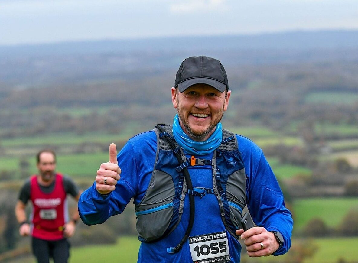 A man running through beautiful countryside giving a thumbs up sign.